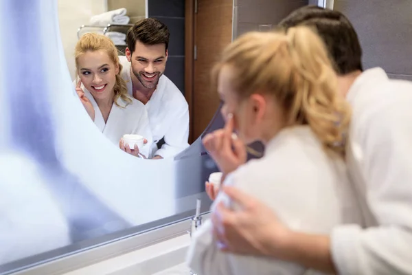 Smiling couple looking at mirror in white bathrobes in bathroom — Stock Photo