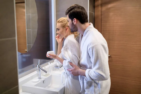 Pareja cariñosa mirando el espejo en batas blancas en el baño - foto de stock