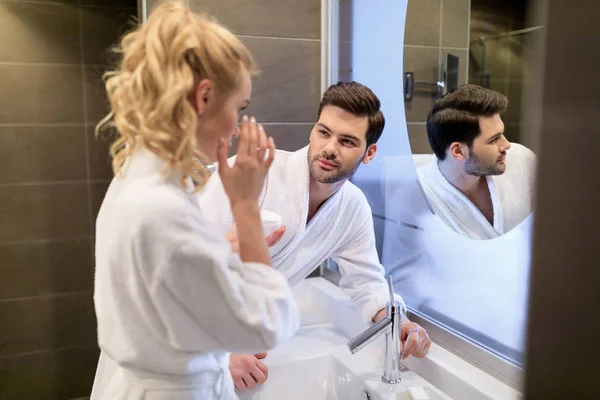 Novio mirando cómo novia aplicando crema en la cara en el baño - foto de stock
