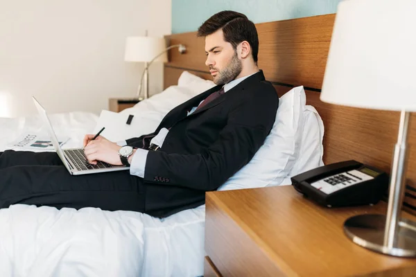 Vista lateral del hombre de negocios acostado en la cama en la habitación de hotel y el uso de ordenador portátil - foto de stock