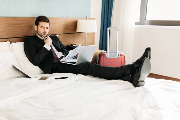 Hombre de negocios acostado en la cama en la habitación de hotel y leyendo el periódico - foto de stock