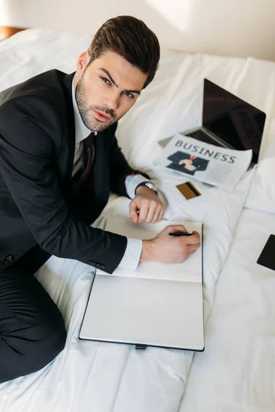 Hombre de negocios acostado en la cama en la habitación de hotel con cuaderno y pluma - foto de stock