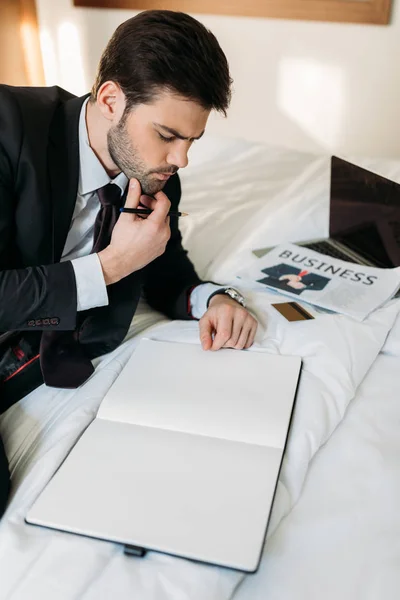 Thoughtful businessman lying on bed in hotel room and looking at notebook — Stock Photo