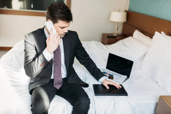 Hombre de negocios guapo hablando por teléfono inteligente y sentado en la cama en la habitación de hotel - foto de stock