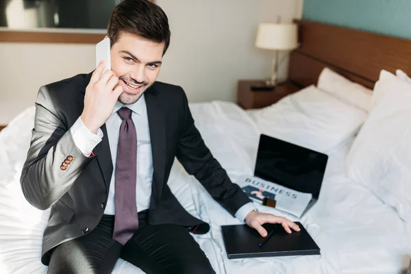 Homem de negócios sorrindo falando por smartphone e sentado na cama no quarto de hotel — Fotografia de Stock