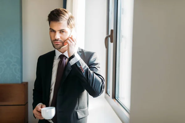 Hombre de negocios guapo hablando por teléfono inteligente en la habitación de hotel y mirando a la cámara - foto de stock