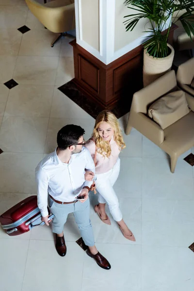 Vista aérea de sonriente pareja de viajeros caminando con equipaje en el hotel - foto de stock