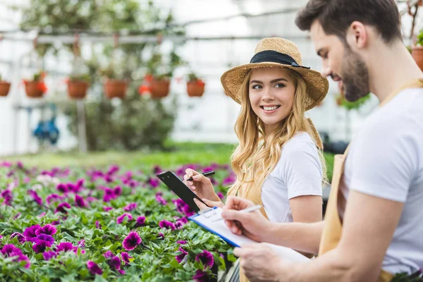 Lächelnde Besitzer von Gewächshäusern, die Klemmbretter an Blumen halten — Stock Photo