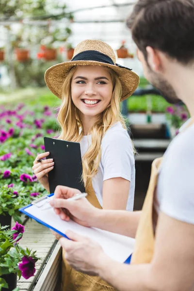 Young male and female gardeners holding clipboards by flowers in glasshouse — Stock Photo