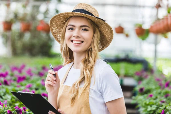 Mujer rubia sosteniendo portapapeles por flores en invernadero - foto de stock