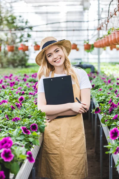 Smiling female gardener holding clipboard by flowers in greenhouse — Stock Photo