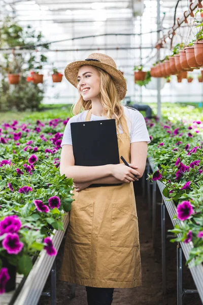 Jardineiro feminino atraente segurando prancheta por flores em estufa — Fotografia de Stock