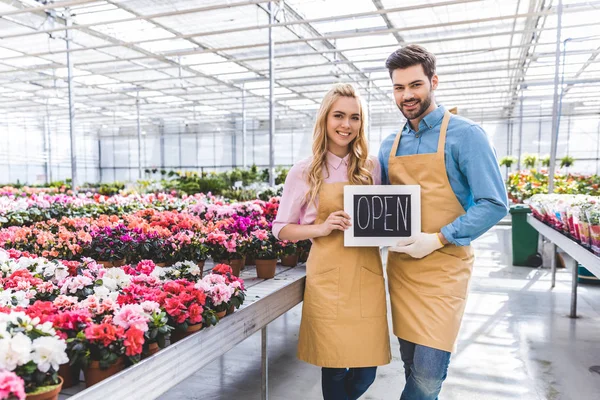 Male and female owners of glasshouse holding Open board by flowers — Stock Photo