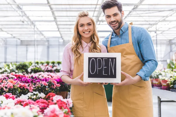 Young man and blonde woman holding Open board by flowers in greenhouse — Stock Photo