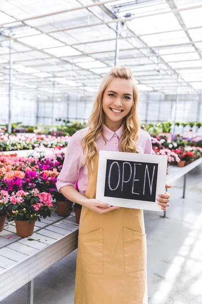 Smiling female owner of glasshouse holding Open board by flowers in greenhouse — Stock Photo