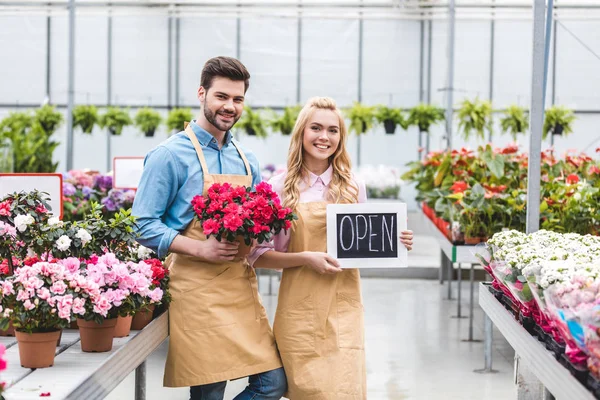 Young man and blonde woman holding Open board by flowers in glasshouse — Stock Photo