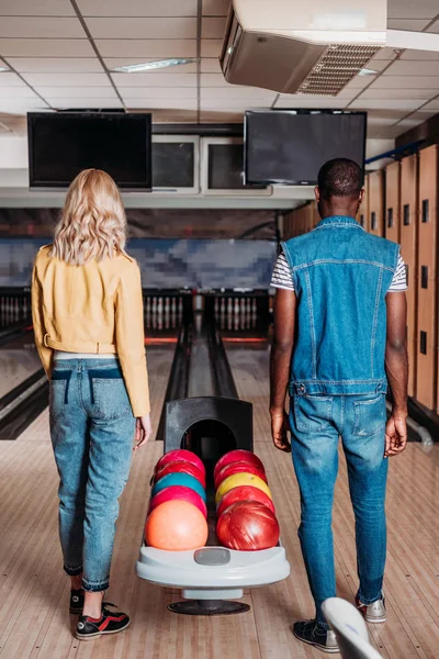 Vista posteriore di coppia multietnica in piedi vicino stand con le palle al bowling club — Foto stock
