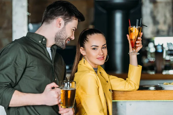 Mujer joven con cóctel mirando a la cámara mientras el hombre coquetea con ella en el bar - foto de stock