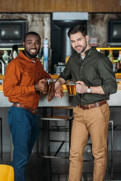 Jovens sorridentes com canecas de cerveja no balcão do bar olhando para a câmera — Fotografia de Stock