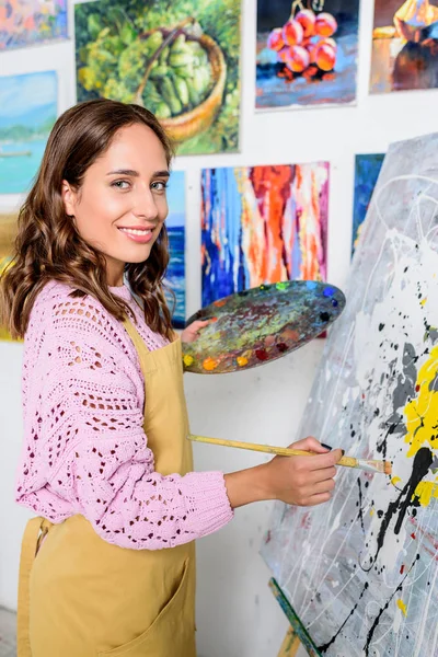 Beautiful female artist painting in workshop and looking at camera — Stock Photo