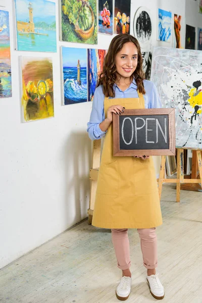 Schöne Künstlerin hält Schild mit geöffnetem Wort in Werkstatt und blickt in Kamera — Stockfoto
