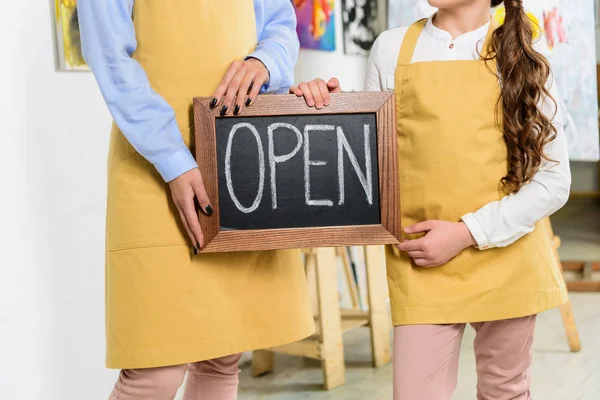 Cropped image of teacher and pupil holding signboard with word open in workshop of art school — Stock Photo