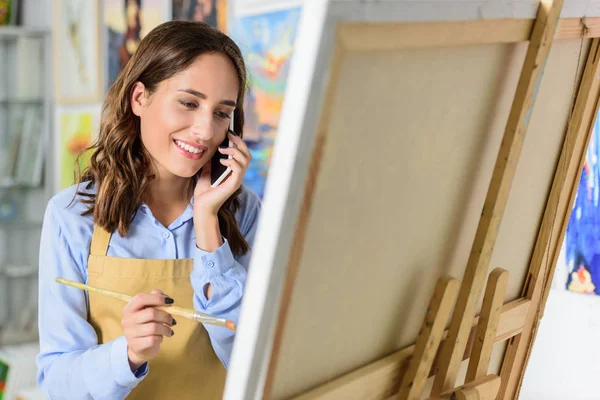 Beautiful female artist talking by smartphone in workshop and painting on canvas — Stock Photo