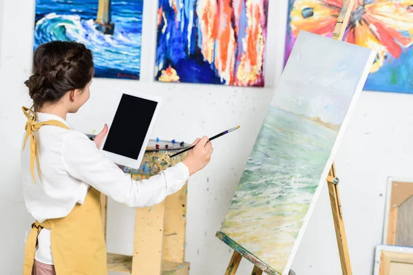 Vista lateral del niño mirando tableta y pintura sobre lienzo en el taller de la escuela de arte - foto de stock