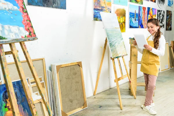 Enfant souriant debout avec tablette près de toile dans l'atelier de l'école d'art — Photo de stock