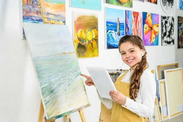 Smiling child standing with tablet in workshop of art school and pointing on canvas — Stock Photo