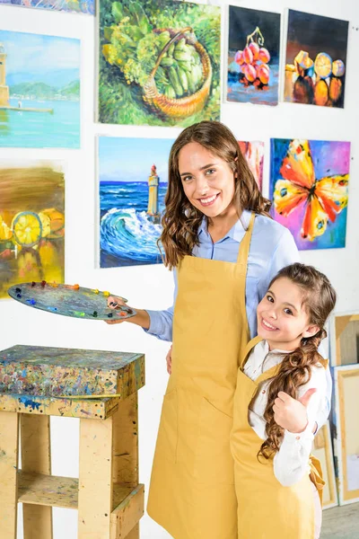 Profesora abrazando pupila y ella mostrando pulgar hacia arriba en taller de arte escuela - foto de stock