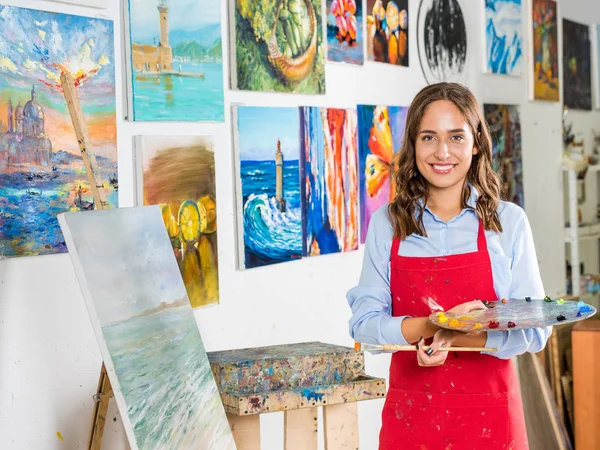 Привлекательная художница держит кисть и палитру рядом с холстом в мастерской — стоковое фото