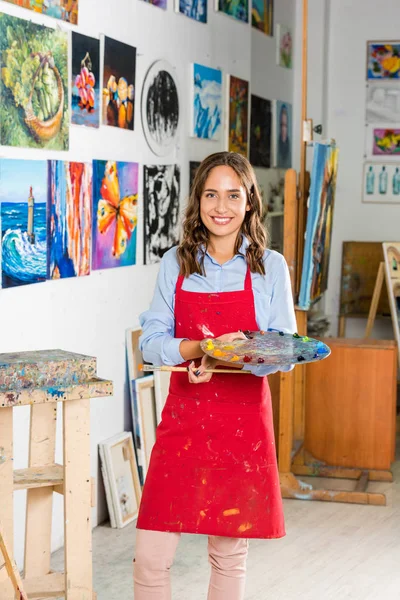 Bella artista femminile che tiene pennello e tavolozza di pittura in laboratorio — Foto stock