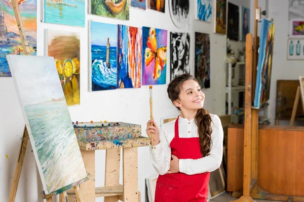 Kid holding painting brush and looking away in workshop of art school — Stock Photo