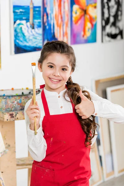 Kid showing thumb up and holding painting brush in workshop of art school — Stock Photo