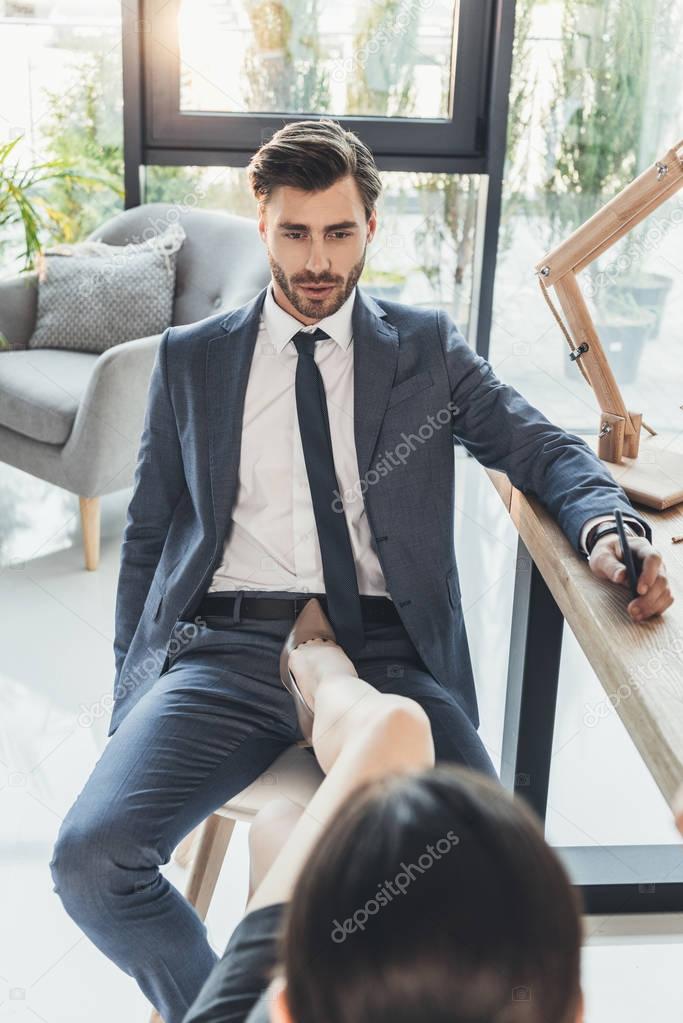 Woman in high heels placing her foot on a chair between legs of young man in business sui