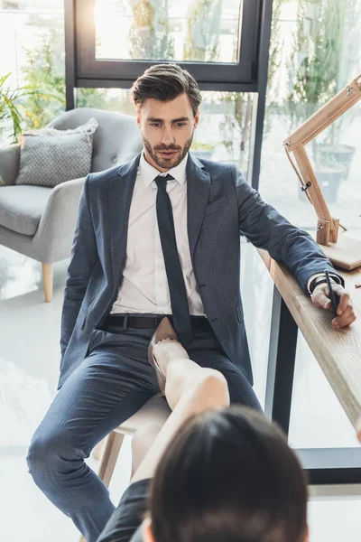 Frau in High Heels setzt ihren Fuß auf einen Stuhl zwischen den Beinen eines jungen Mannes in Business-Uniform — Stockfoto