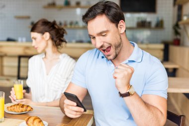 Onun smartphone ve arka plan üzerinde oturan mutsuz kadın kullanan bağımlısı adam. Adam kutlamak kazanmak kumar mobil uygulama futbol bahis, bahis spor kumar