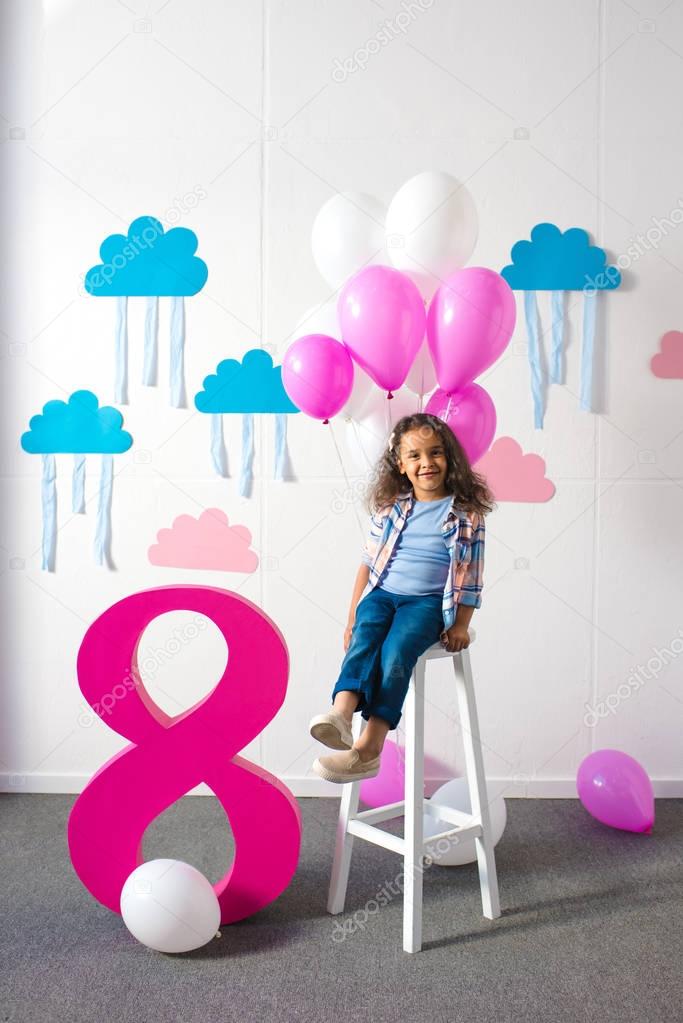 Adorable african american girl sitting on stool with balloons and smiling at camera at birthday party