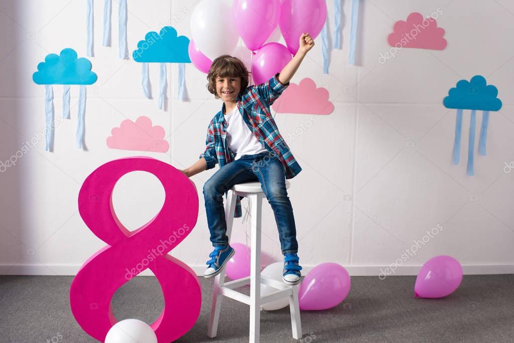 Happy little boy sitting on stool with balloons and smiling at camera at birthday party