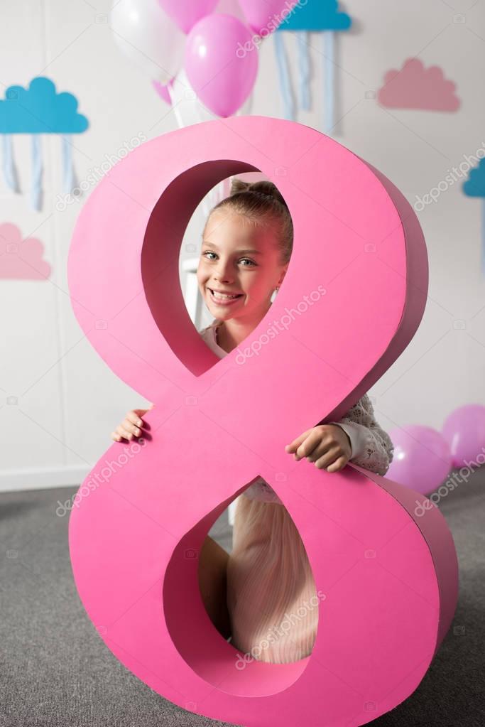 Adorable little girl holding big pink decorative number eight and smiling at camera at birthday party
