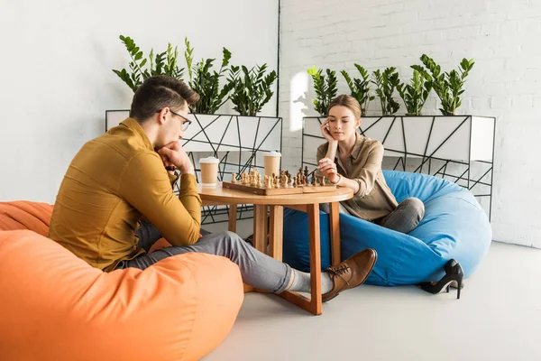 Молодая целеустремленная пара играет в шахматы, сидя на мешках — стоковое изображение Молодая Целеустремленная Пара Играет Шахматы Сидя Мешках — стоковое фото