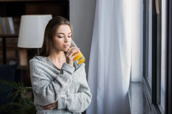 Bella ragazza che beve succo d'arancia vicino alla finestra al mattino — Foto stock