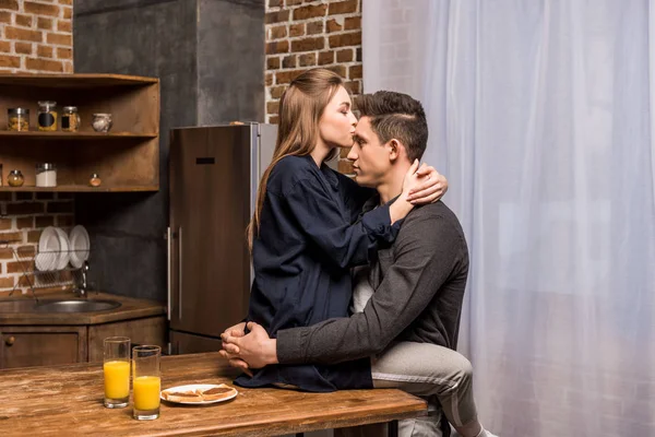 Freundin sitzt auf Tisch in Küche und küsst Freund auf die Stirn — Stockfoto