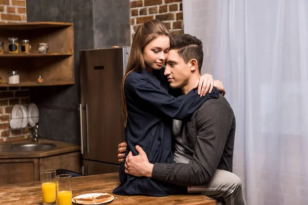 Novia sentada en la mesa en la cocina y abrazando novio - foto de stock