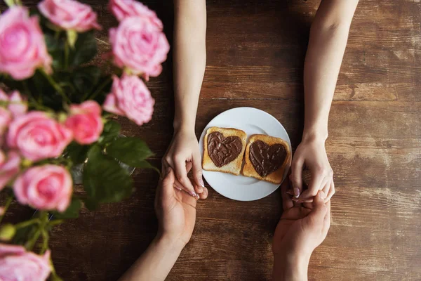 Imagen recortada de pareja cogida de la mano cerca de tostadas con pasta de chocolate en forma de corazones, concepto de día de San Valentín - foto de stock