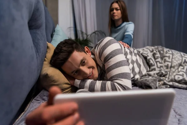 Fidanzato sorridente guardando qualcosa su tablet e fidanzata seduto arrabbiato sul letto — Foto stock