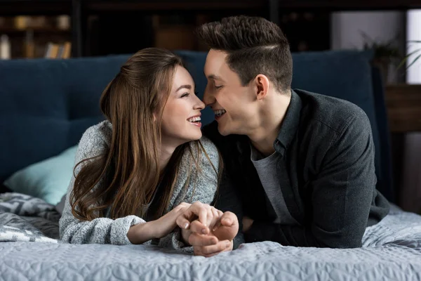 Sonriente pareja tocando con narices y tomándose de la mano en la cama - foto de stock