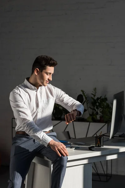 Handsome young businessman sitting on work desk and looking at watch — Stock Photo