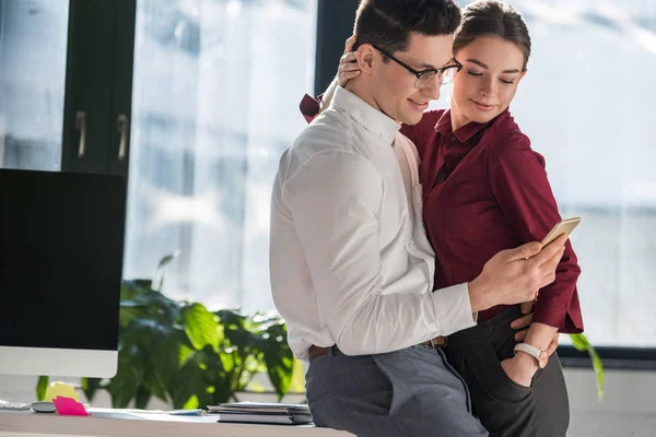 Colegas abrazando y utilizando el teléfono inteligente juntos en la oficina - foto de stock